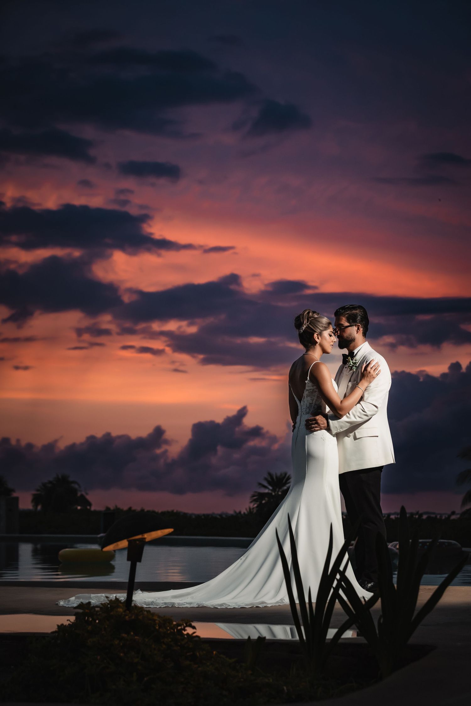 Wedding couple embraces on dock with dramatic purple and orange sunset sky.