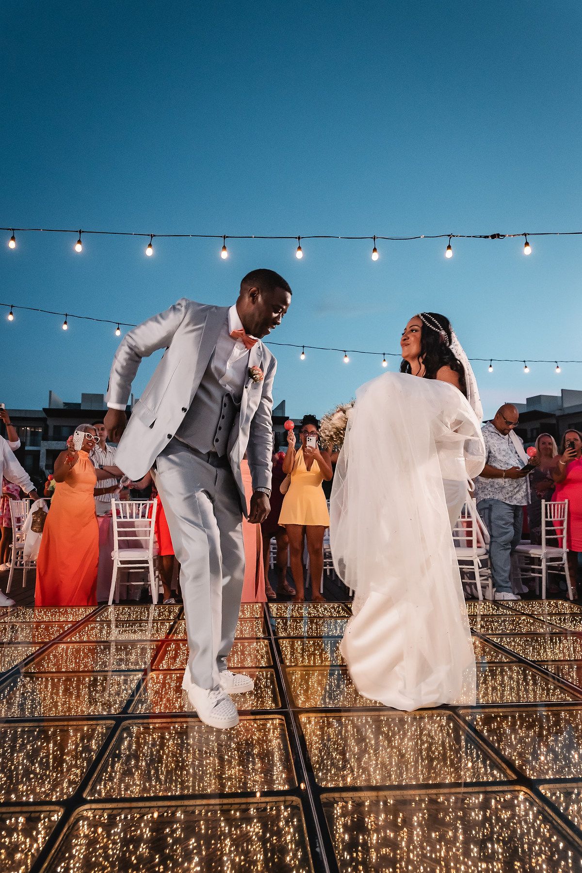 A wedding couple and guests celebrate with string lights on a rooftop terrace during sunset.