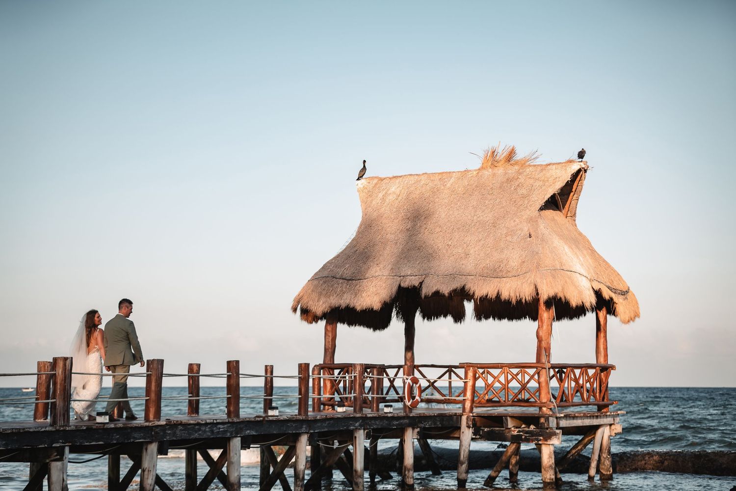 Thatched-roof pavilions on a wooden pier extending over ocean waters at sunset.