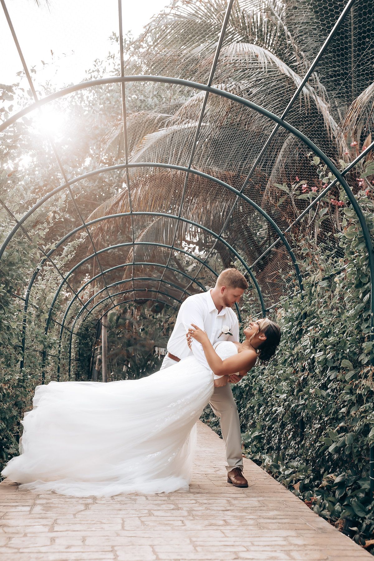 A romantic wedding couple shares an intimate dip pose in a garden tunnel pathway.