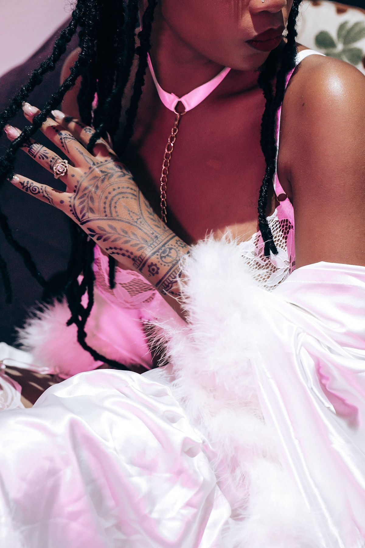 Close-up artistic photo showing hands with intricate henna tattoos against pink feathered fabric in dramatic lighting.