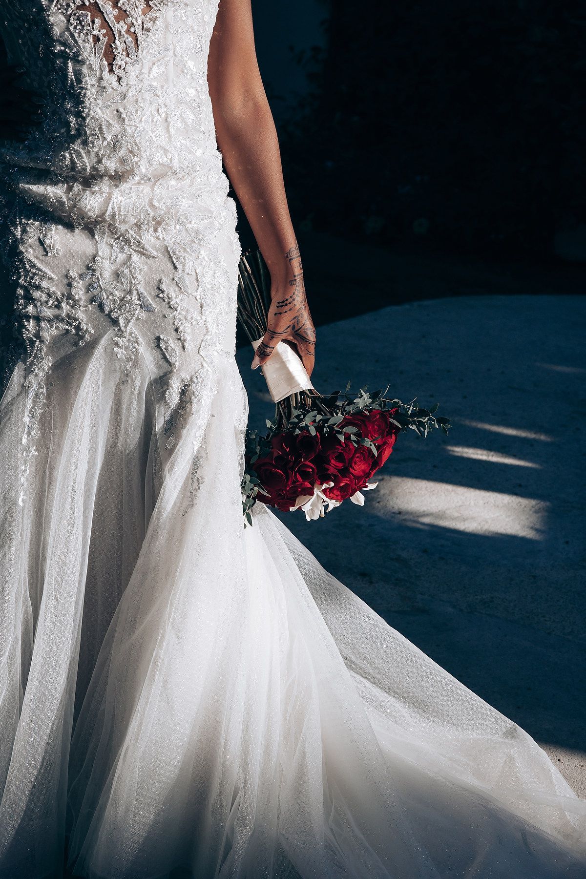 Close-up details of a white lace wedding dress with flowing tulle skirt and deep red bouquet against dark background.