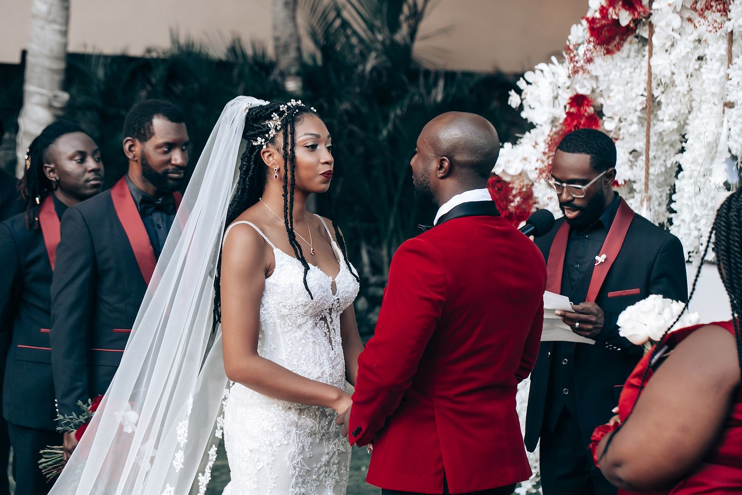 A wedding party poses together wearing elegant red and black formal attire with white decor in the background.
