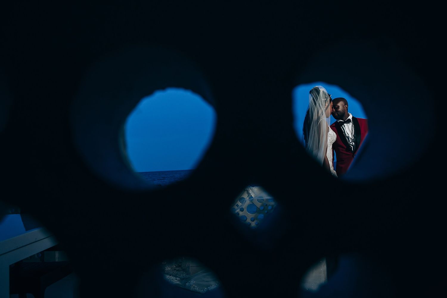Artistic wedding photo captured through circular holes showing couple in blue silhouette against dark background.