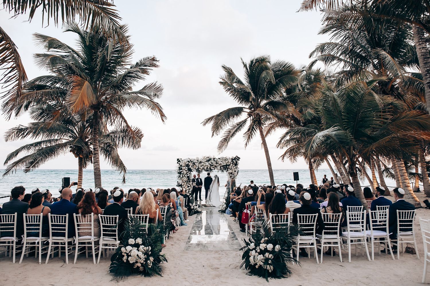 Beach wedding venue with white chairs arranged in rows under palm trees.
