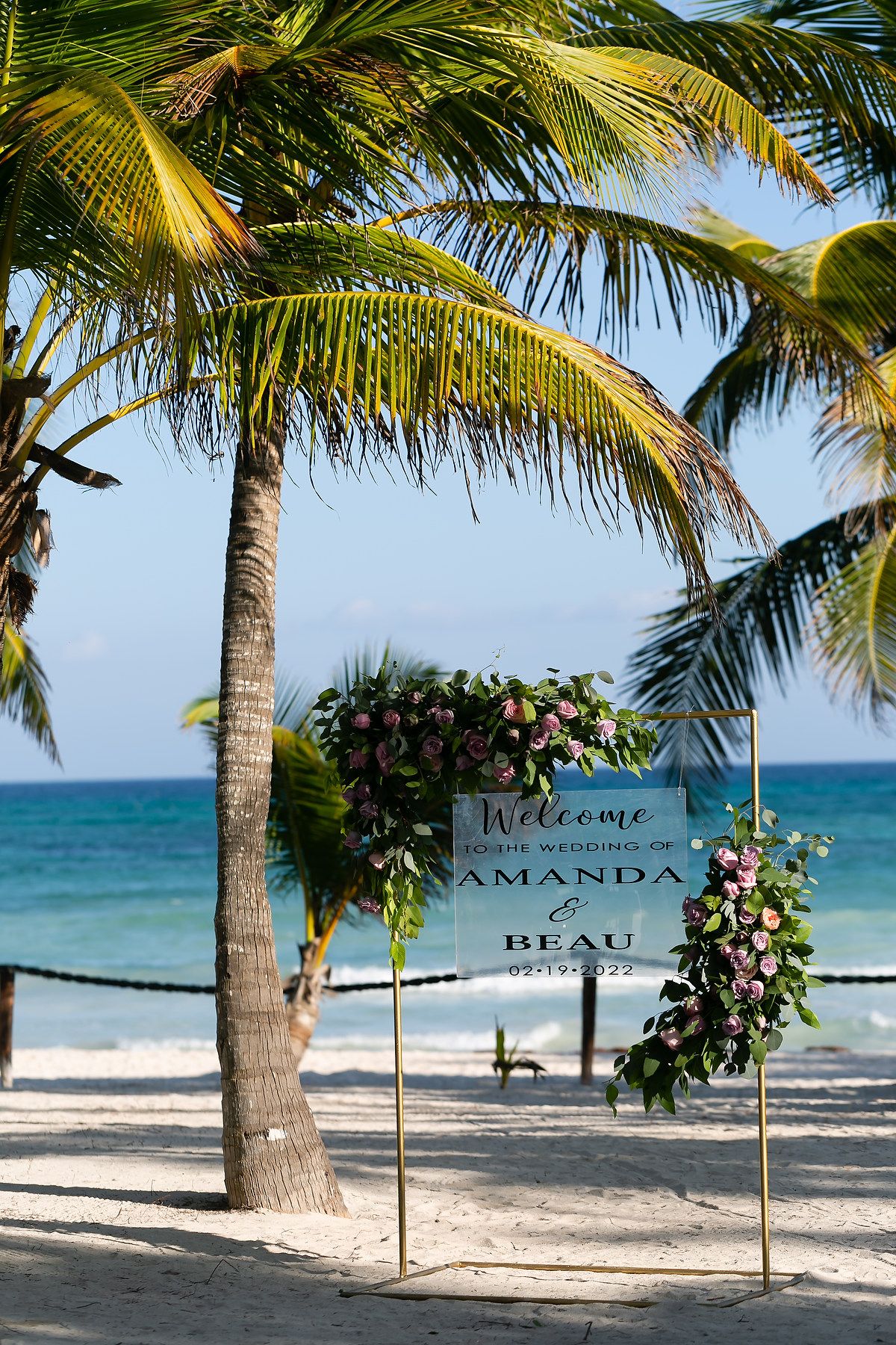 Welcome signs for a beach wedding line a sandy path between palm trees with turquoise ocean views.