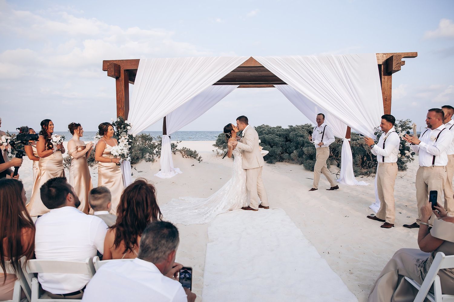 Beachfront wedding ceremony with white canopy and guests seated on white chairs.