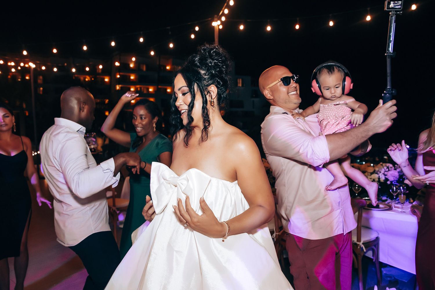 Guests dancing and celebrating at a dimly lit reception with string lights and purple accents.