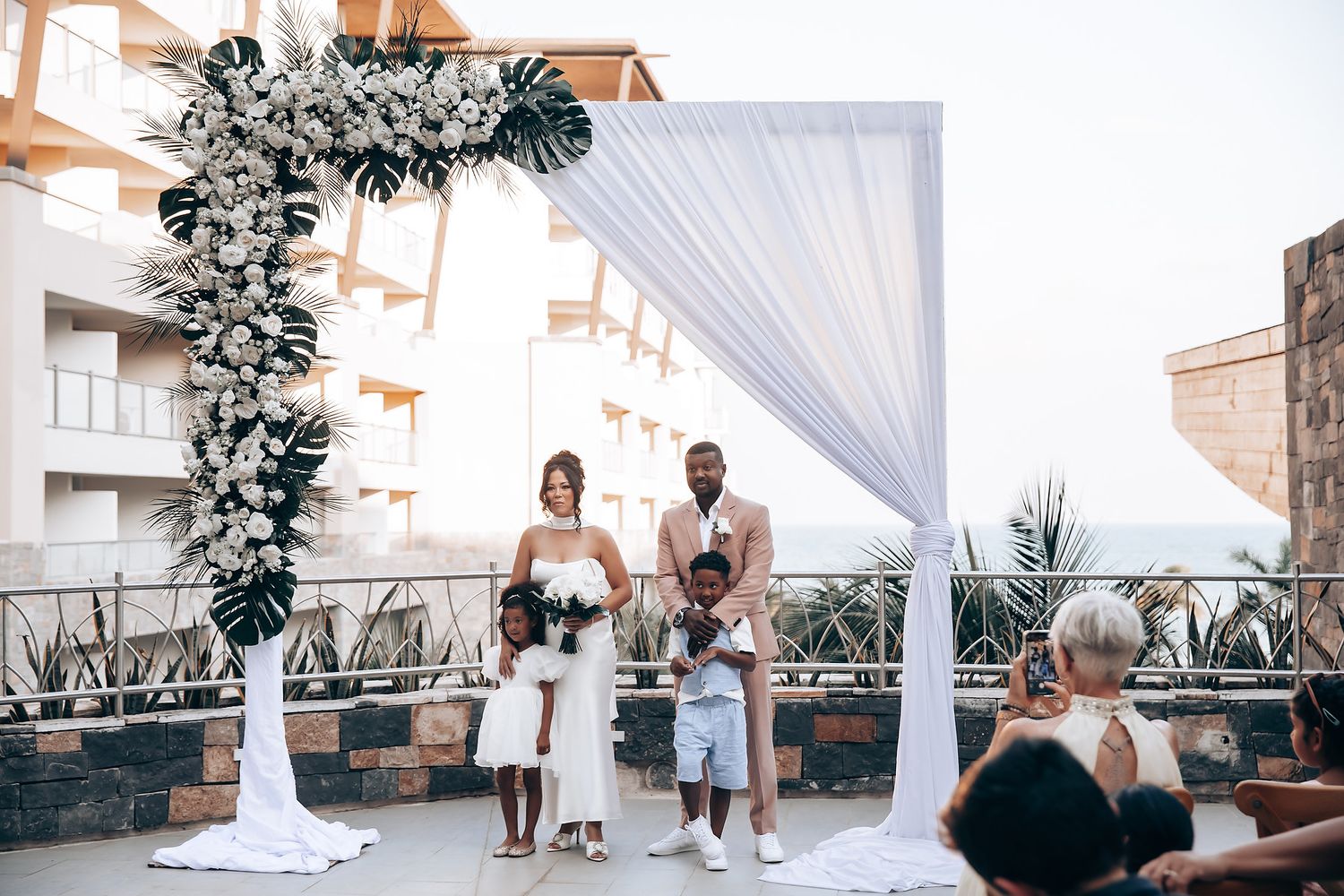 Outdoor wedding ceremony setup with white draping and floral arch against a coastal backdrop.