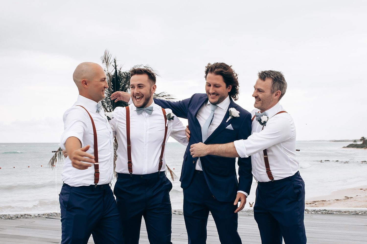 Groomsmen in suspenders and navy pants share a laugh on a cloudy beach.