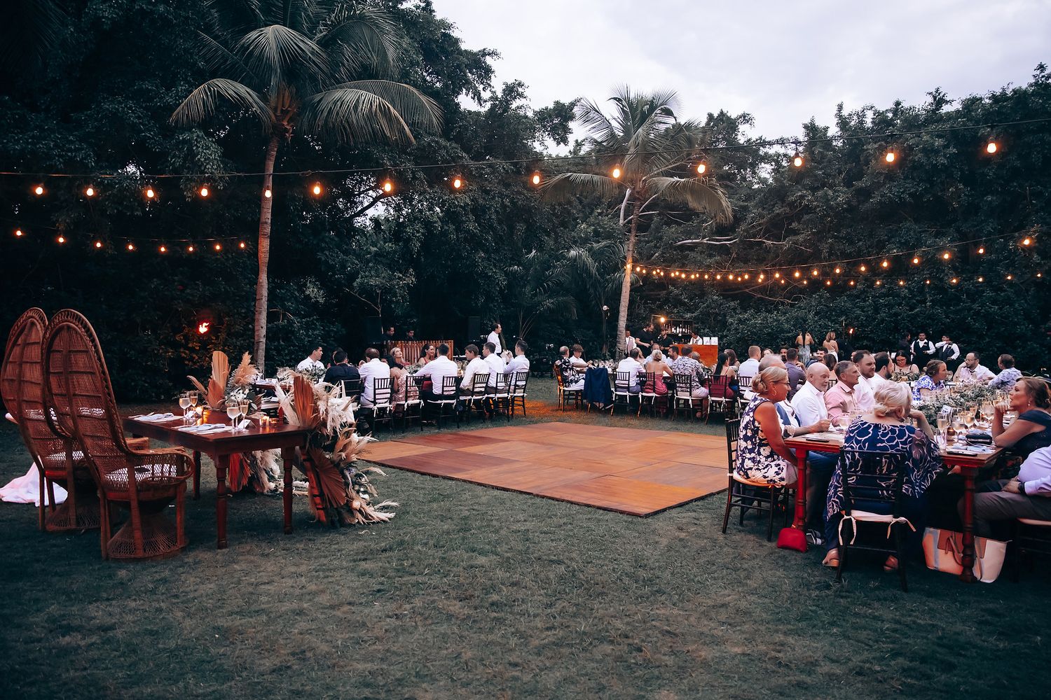 Outdoor wedding reception setup with string lights and wooden dance floor surrounded by tropical landscaping.