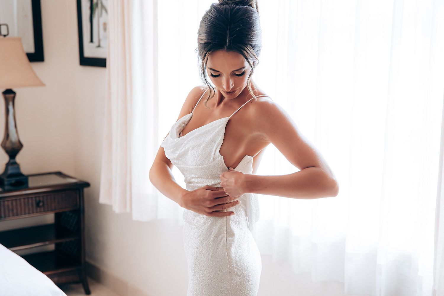 A person in a white dress adjusts their outfit while standing in a sunlit room.