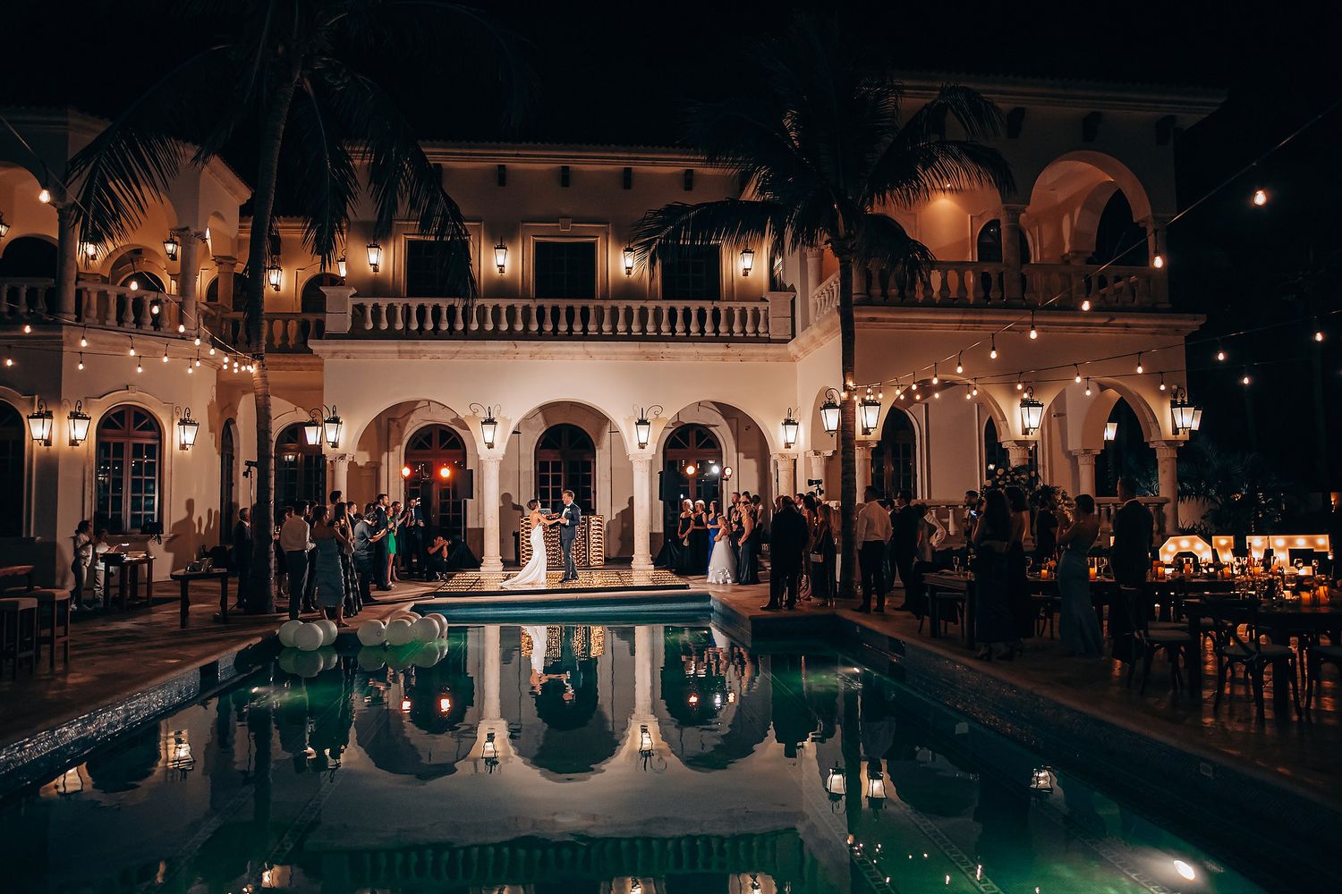 Elegant nighttime wedding reception at a Mediterranean-style venue with illuminated pool and arched architecture.