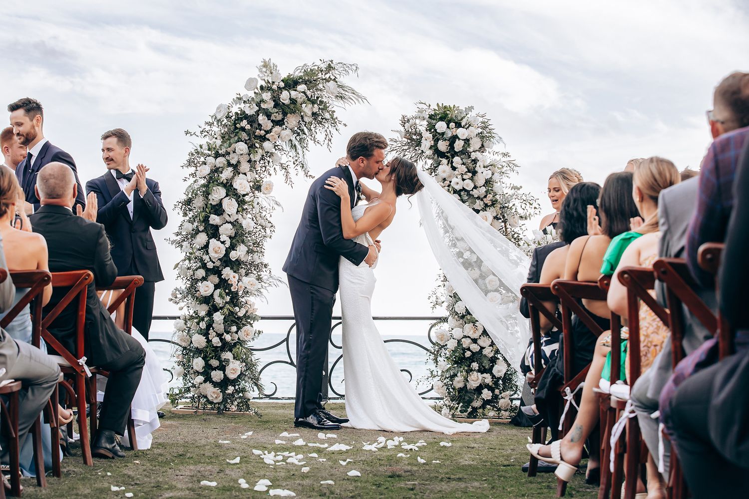 Panoramic view of an outdoor wedding ceremony with guests seated under palm trees and cloudy skies.