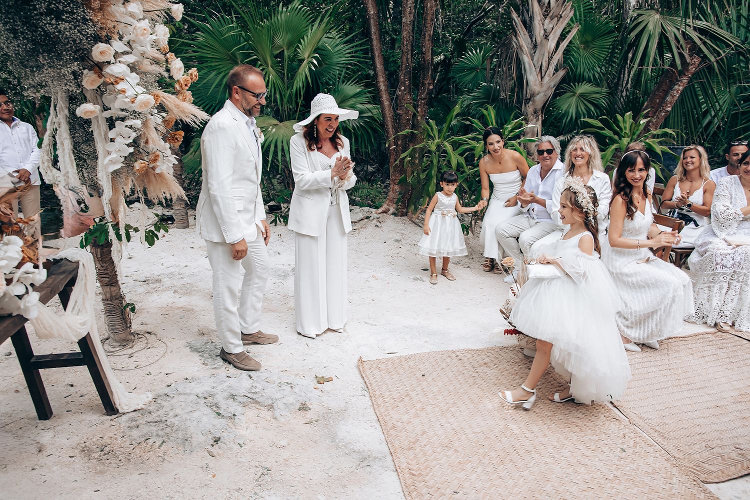 Wedding guests dressed in all white gather on a tropical beach path surrounded by palm trees and lush greenery.