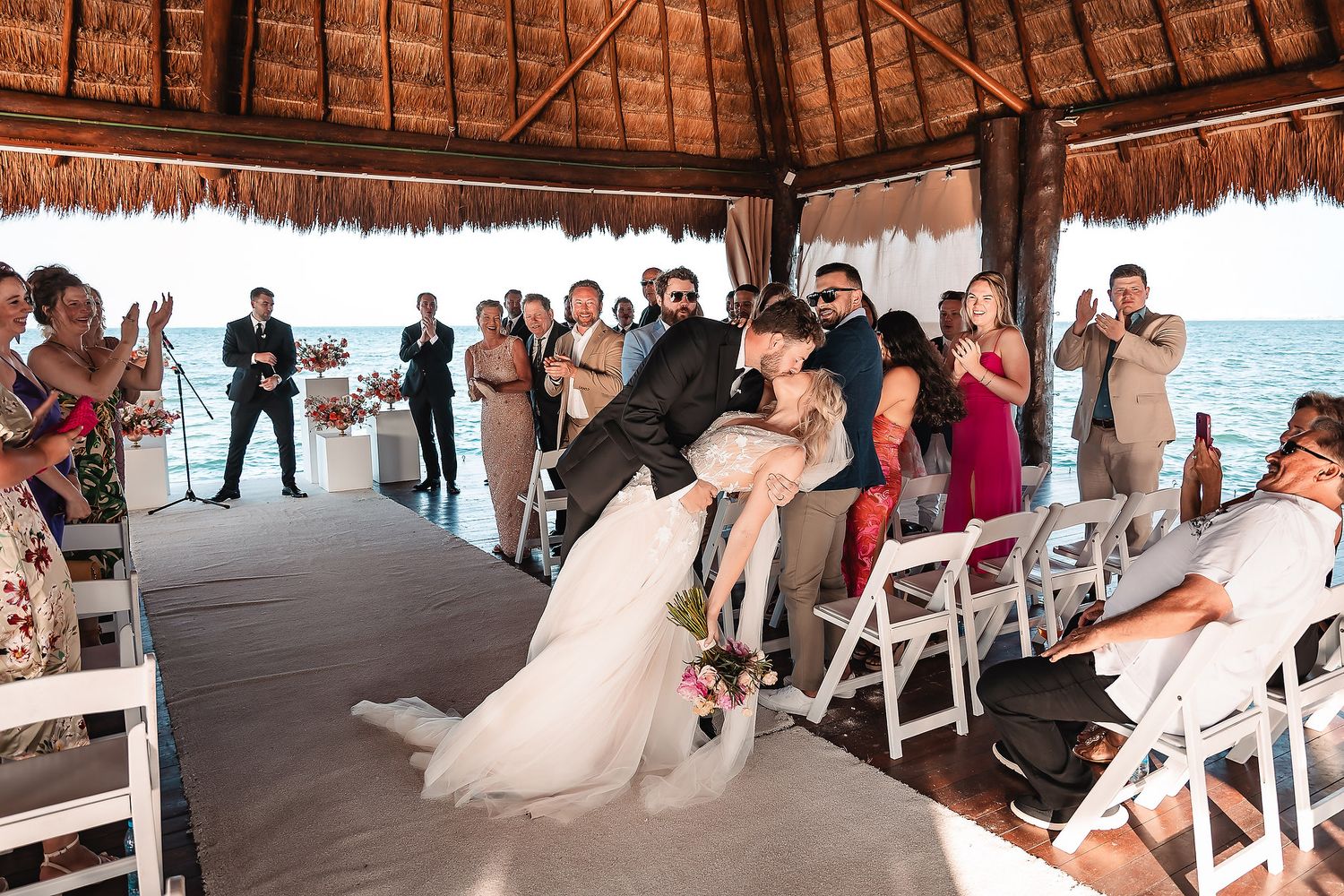 Wedding guests watch ceremony from white chairs under rustic thatched roof venue.