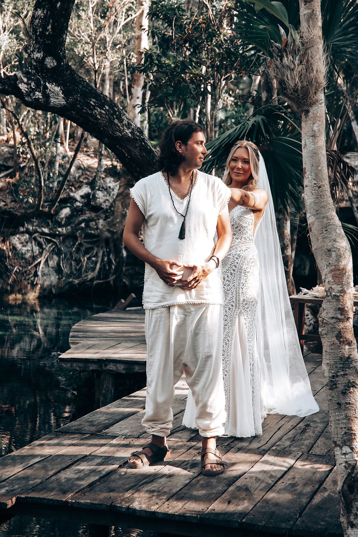 People in white clothing sitting on a wooden deck surrounded by trees during an outdoor ceremony.