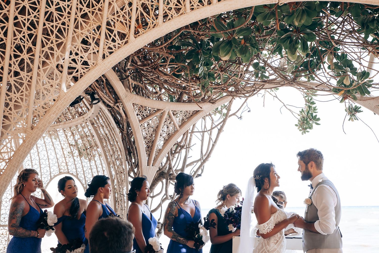 Guests stand in a semicircle under a decorative white pergola during a beach wedding ceremony.