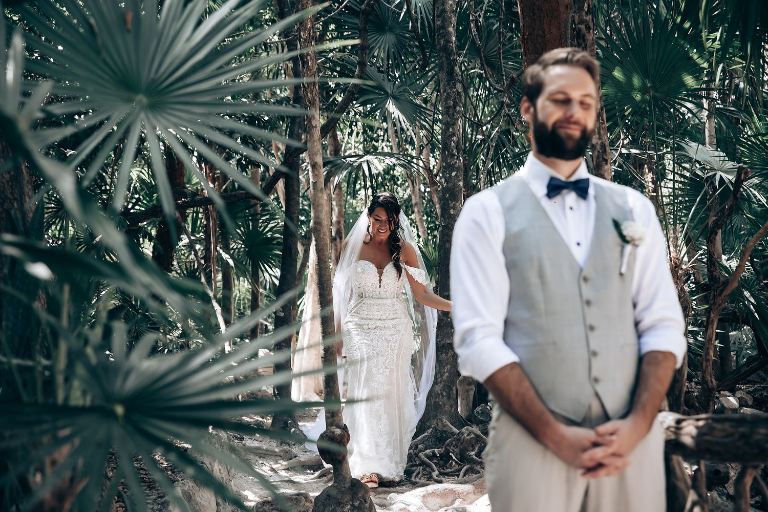 Wedding party members in light gray vests and white shirts pose together in a tropical palm setting.