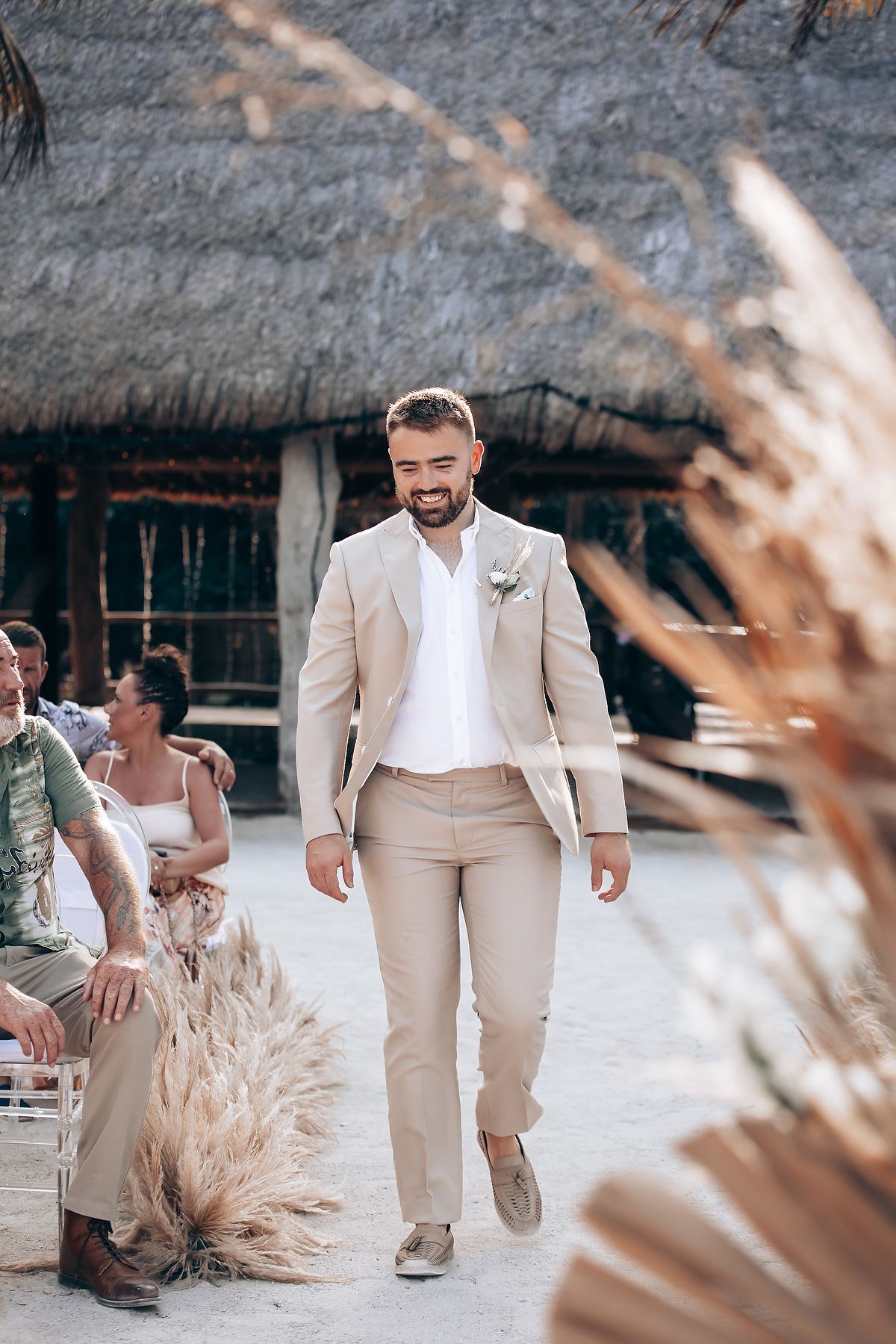 Wedding guests in beige and white attire walk along a beach path with dried pampas grass decor.
