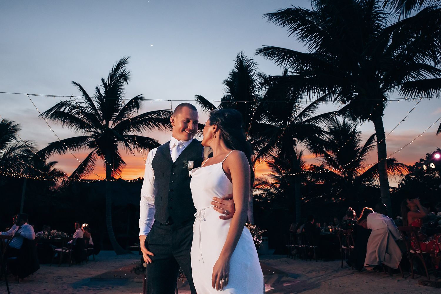Wedding couple poses against a stunning tropical sunset with palm trees silhouetted in the background.