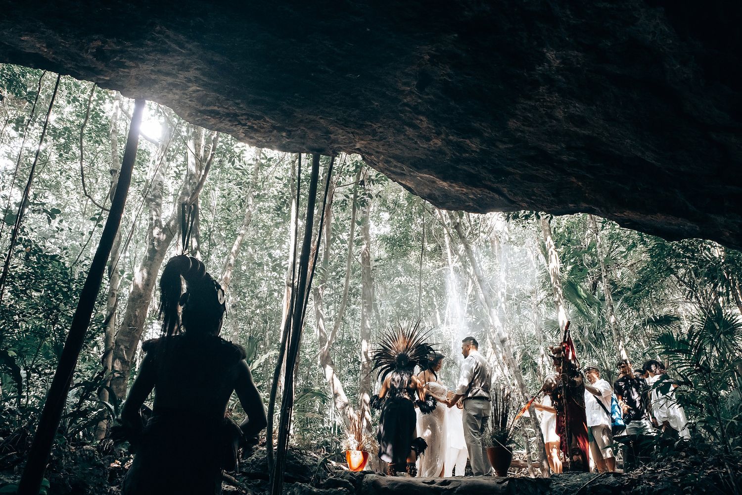 A wedding ceremony takes place in a dramatic cave setting with natural light filtering through trees in the background.