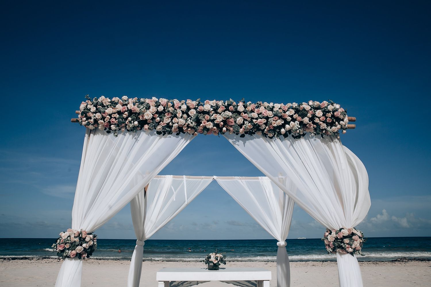 White beach wedding canopy with floral decorations against blue ocean backdrop.