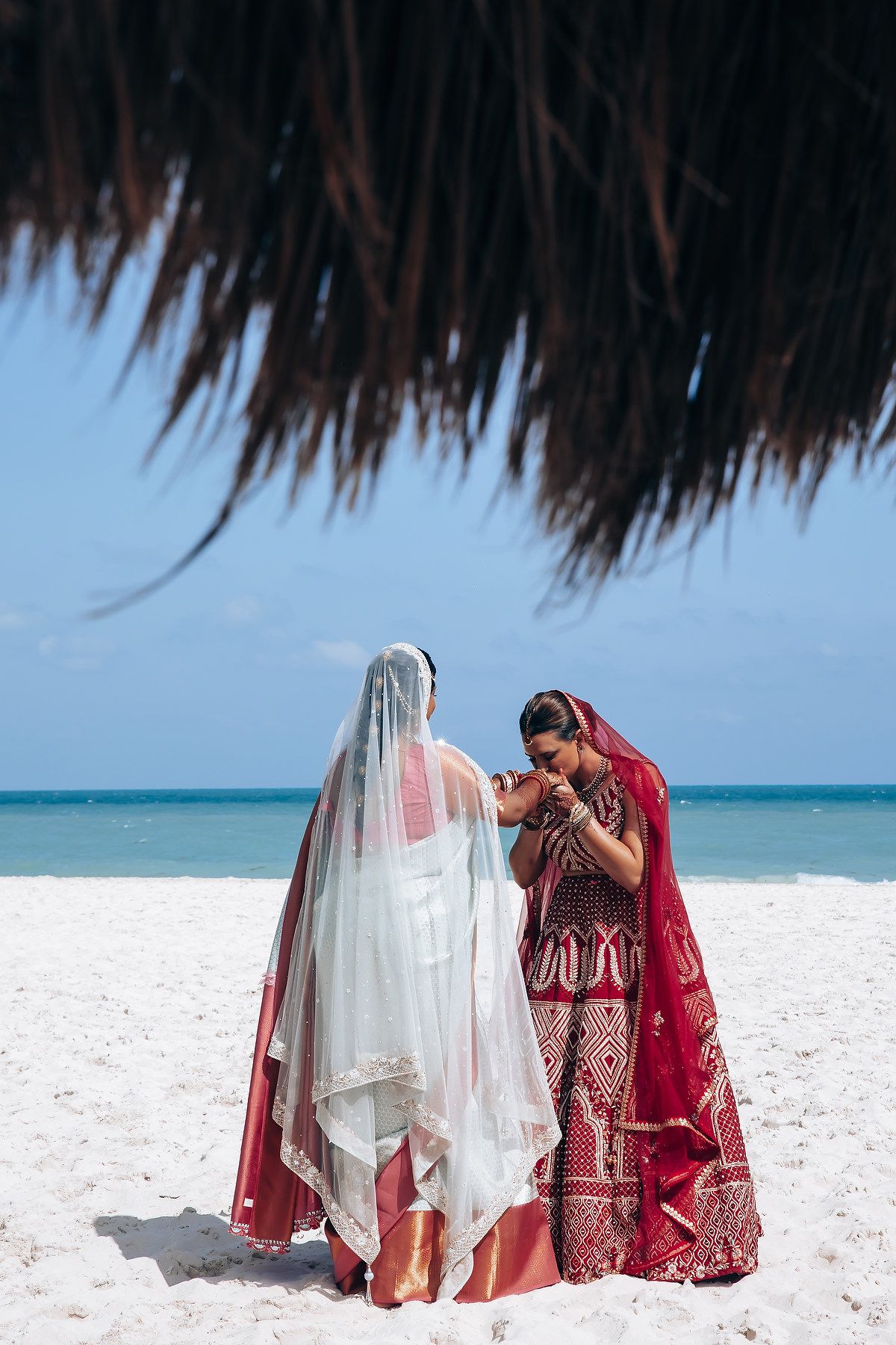 Two people in traditional wedding attire stand together on a white sandy beach under a thatched palapa.