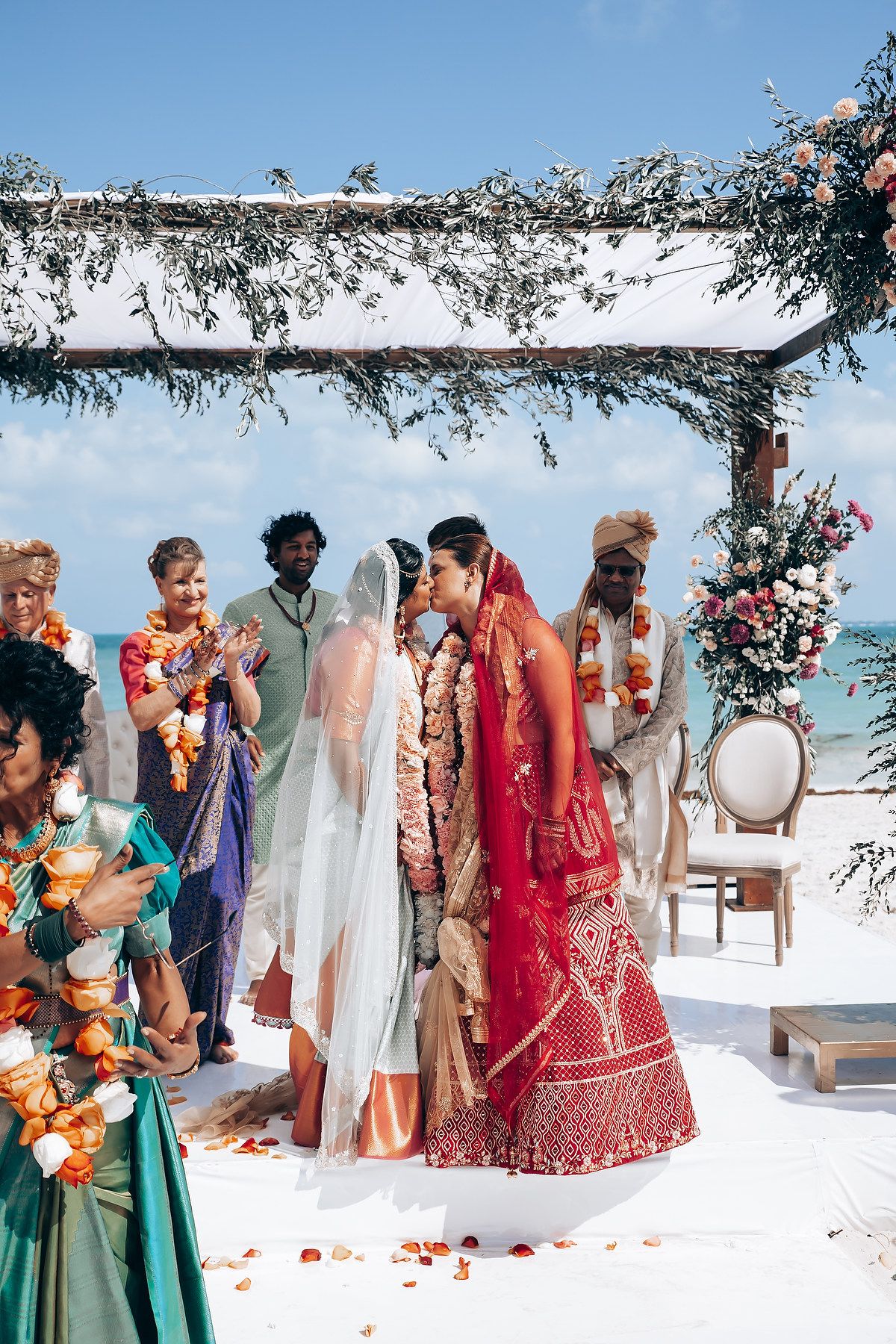 Beachside wedding ceremony with guests in colorful attire under a floral-decorated arch.