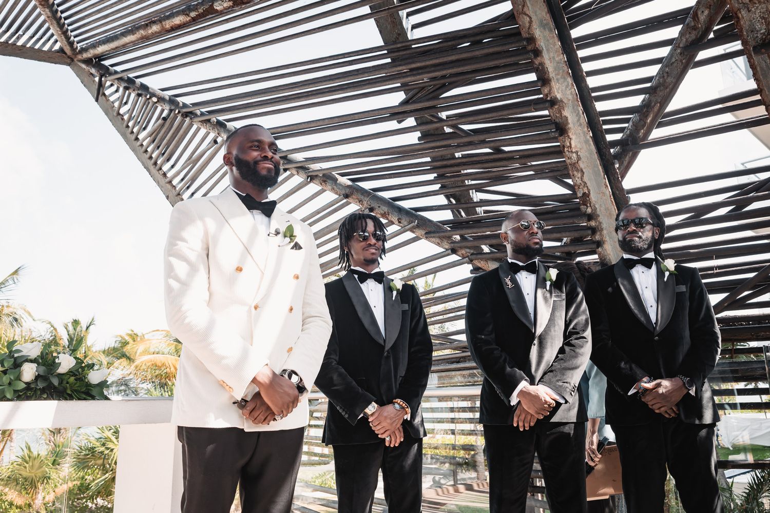Groom in white tuxedo stands with groomsmen in black suits under wooden structure.