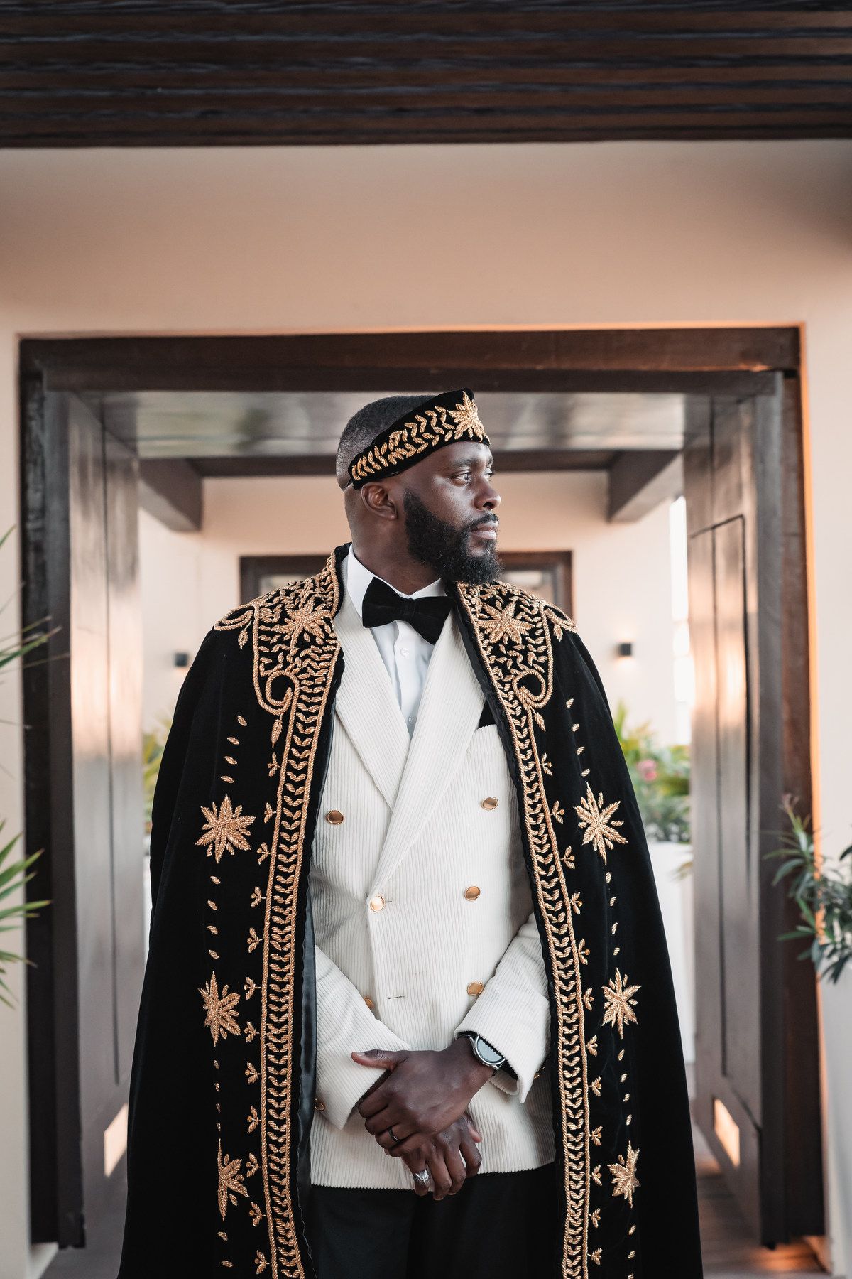 Groom wearing elegant black and gold embroidered cape with matching crown at a luxury wedding ceremony.