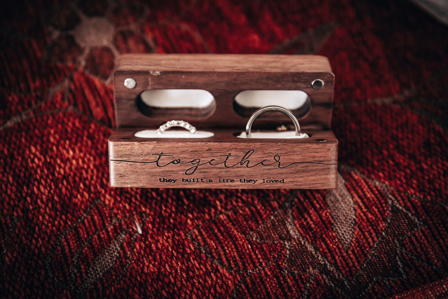 Wooden wedding ring box engraved with together holding two elegant rings on a red textured surface.