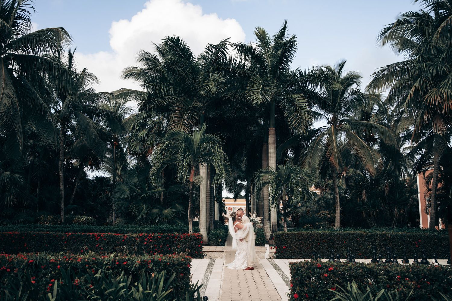 A bride in a white lace gown stands on a wooden path surrounded by tall tropical palm trees at Dreams Tulum
