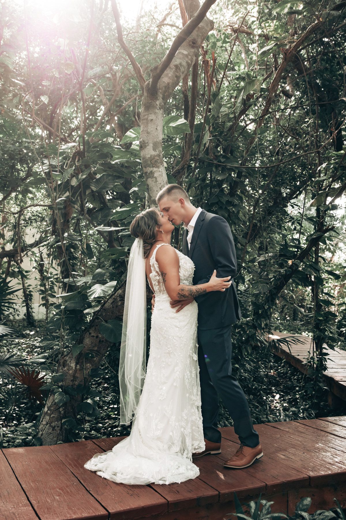Bride and groom sharing a romantic kiss on a wooden deck surrounded by lush tropical greenery on their wedding day.