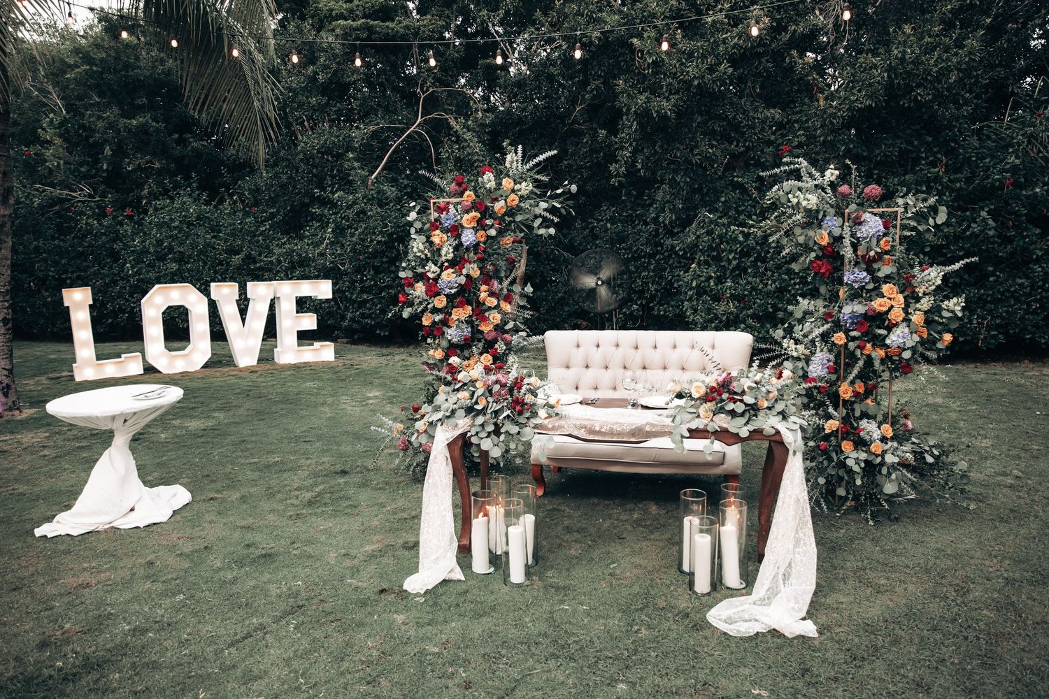 Outdoor wedding sweetheart table setup with lush floral arrangements, candles, and illuminated LOVE sign.