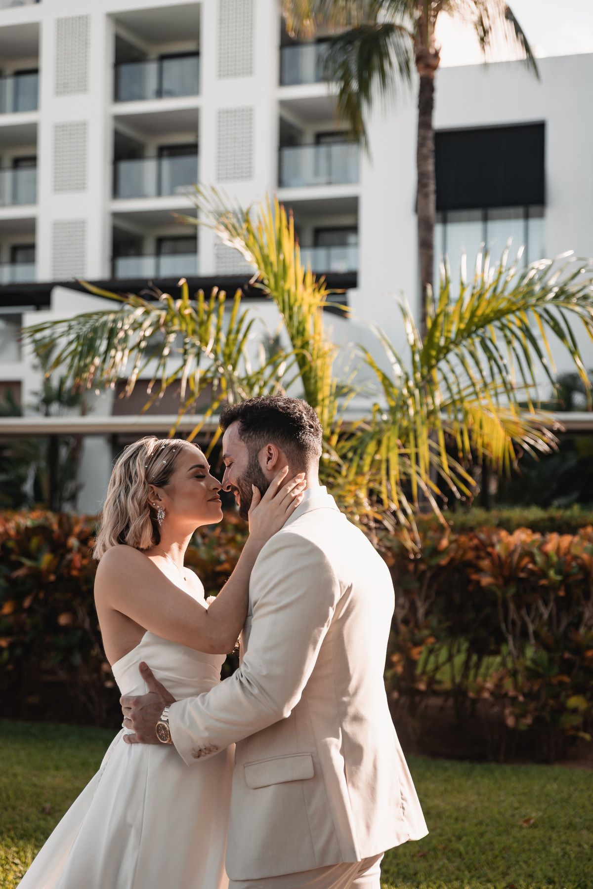 Bride and groom sharing romantic kiss in front of tropical resort with lush palm trees.