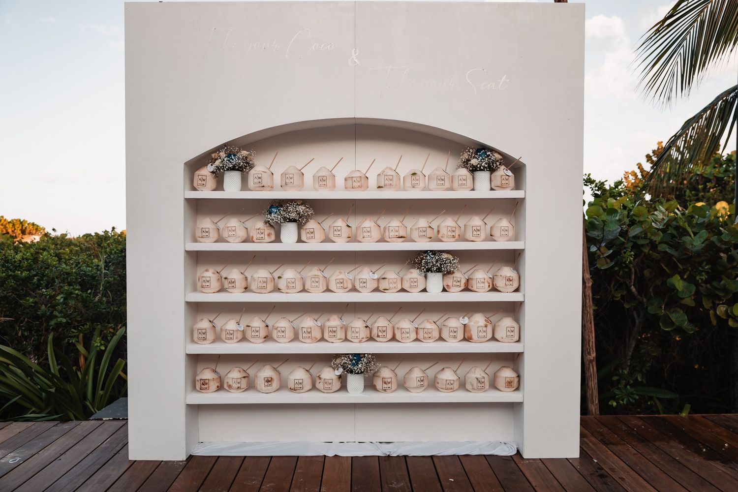 White arched display shelf outdoors holding rows of small wedding favor boxes and mini potted plants.