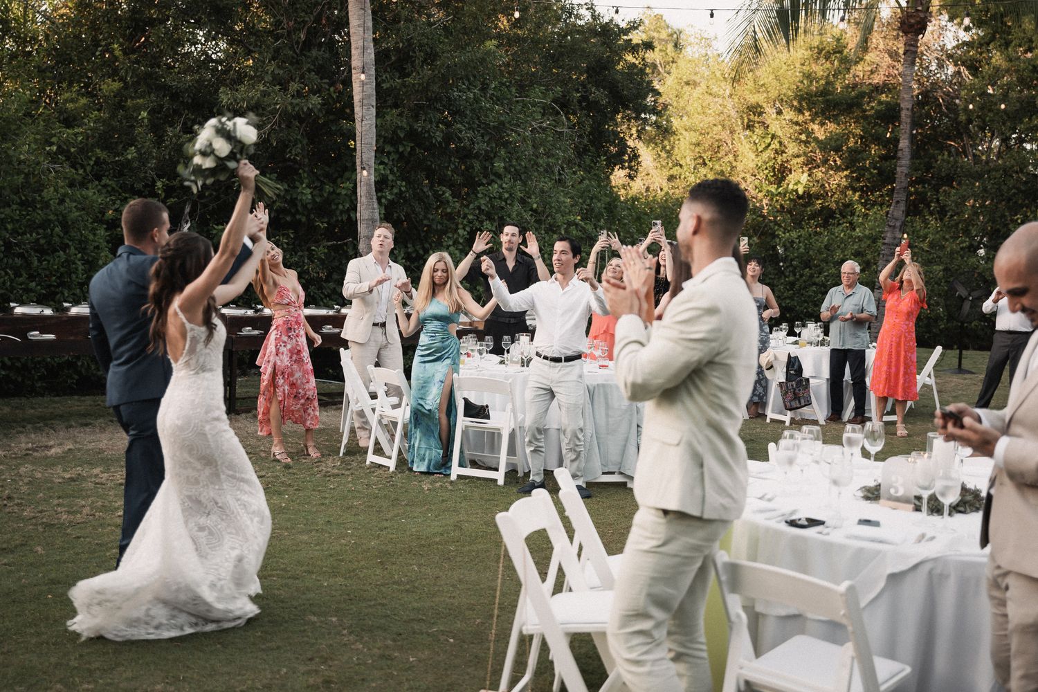 Bride tosses bouquet to excited wedding guests gathered on lawn at outdoor garden wedding reception.