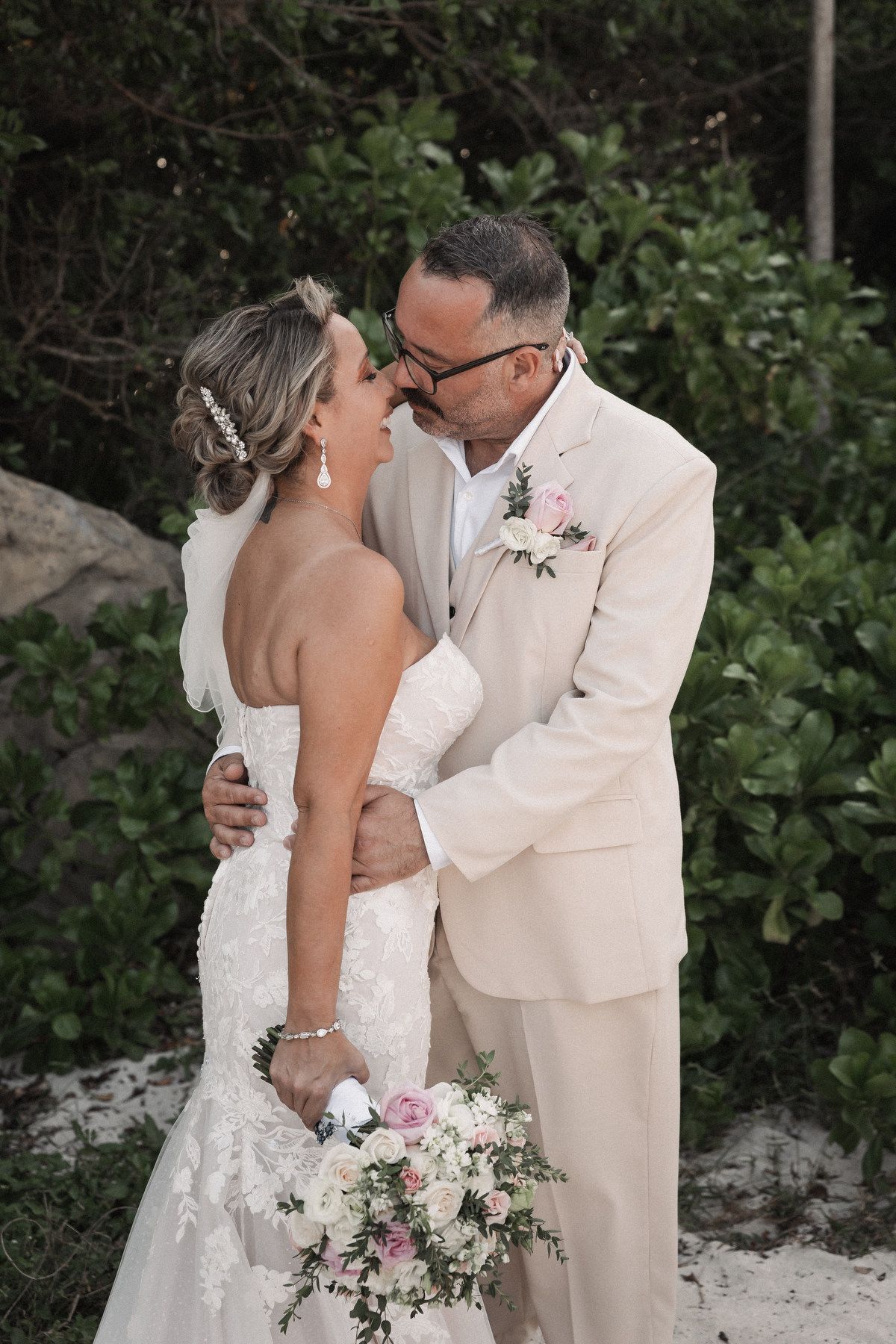 Bride and groom share a romantic forehead touch, she holds pink bouquet, both in white wedding attire outdoors.
