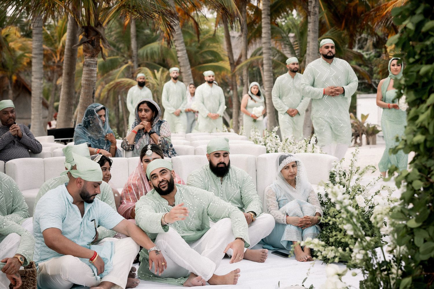 Wedding guests in mint green kurtas gather at outdoor Sikh ceremony surrounded by palm trees.