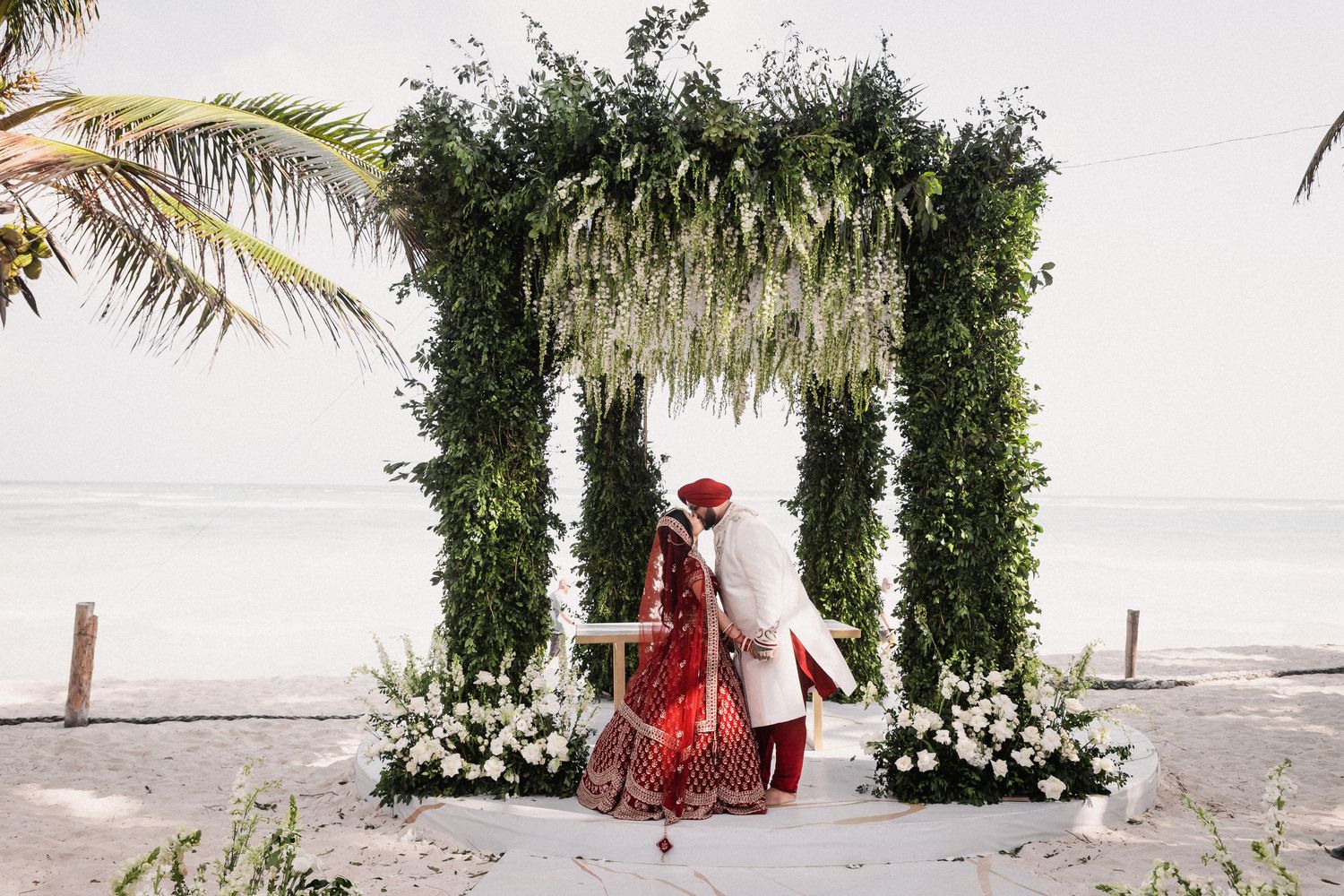 Couple exchanges vows under lush green floral arch on a tropical beach during an Indian wedding ceremony.