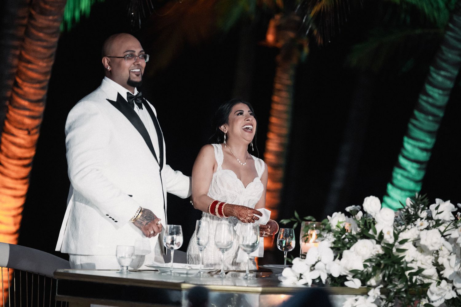 Smiling bride and groom in white tuxedo stand at floral decorated sweetheart table at tropical wedding.