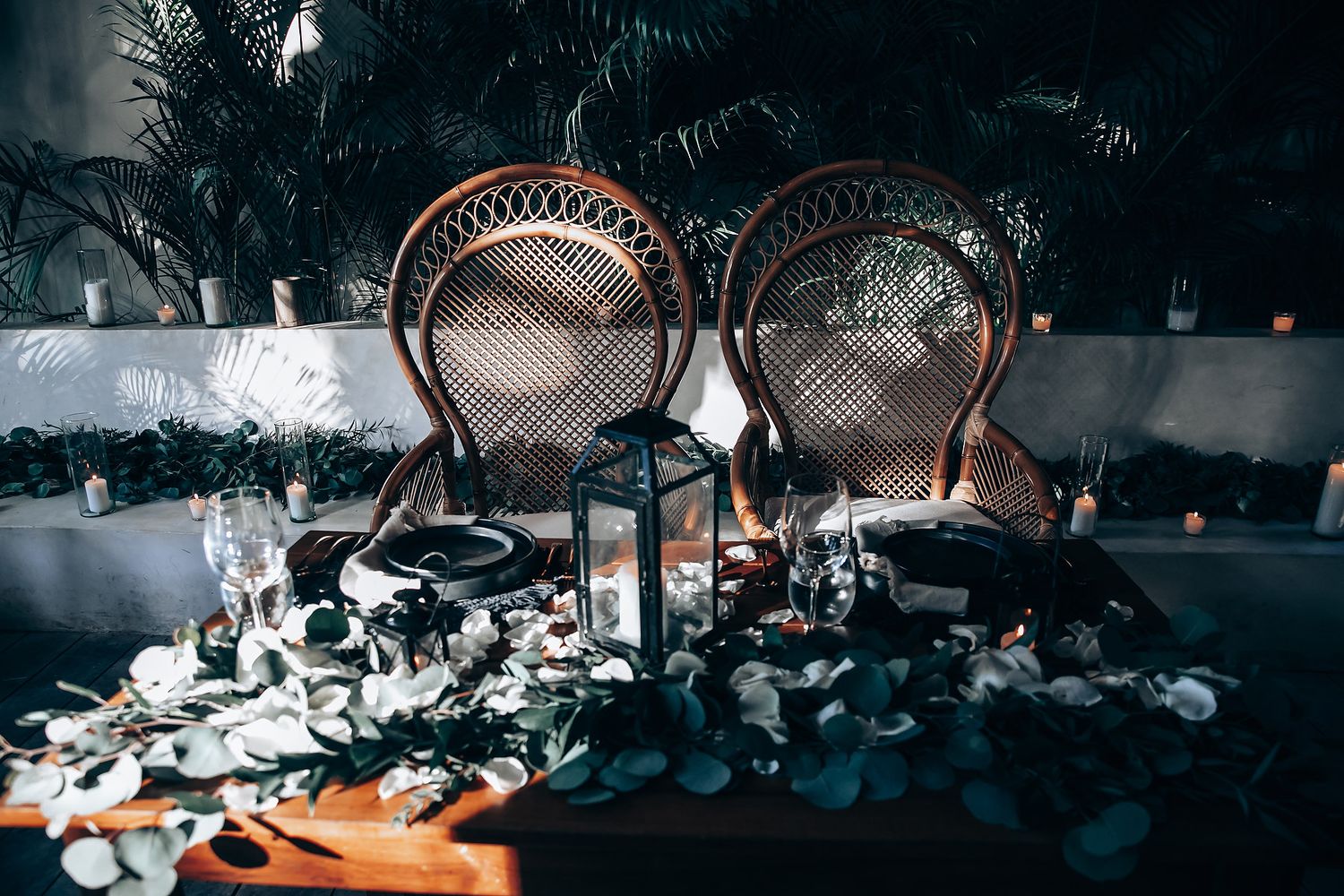 Two vintage peacock chairs surrounded by scattered rose petals and lanterns at an evening reception.