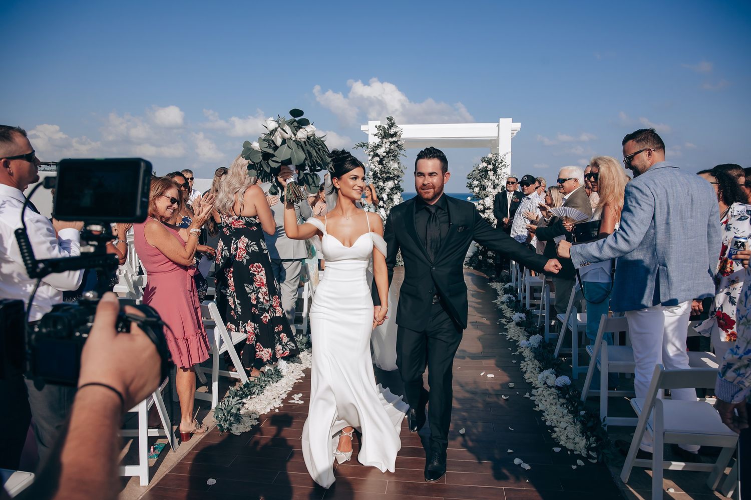 Wedding guests seated outdoors at a ceremony with blue sky and white chairs.