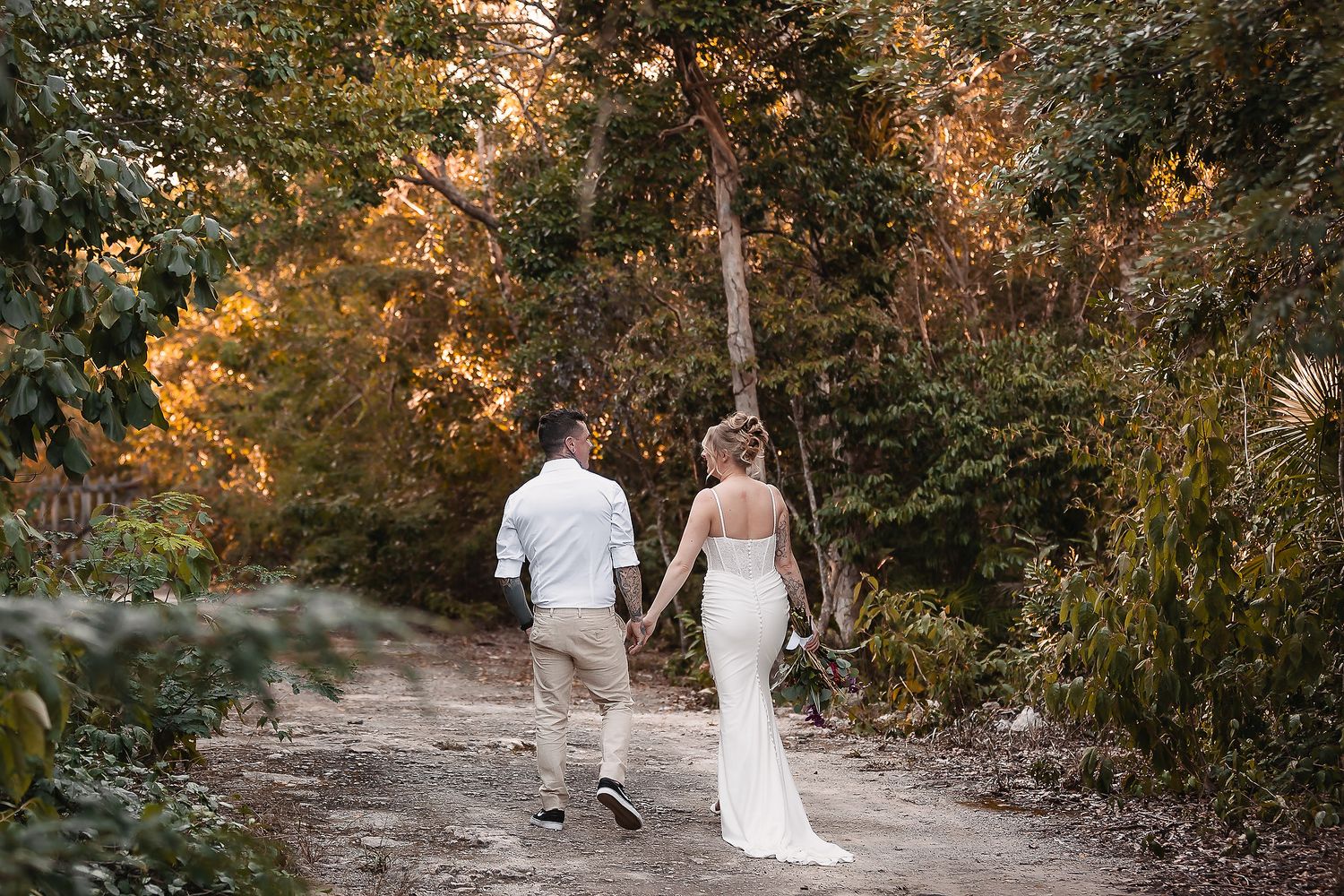 Bride and groom walking hand in hand down a dirt path surrounded by lush tropical greenery at golden hour.
