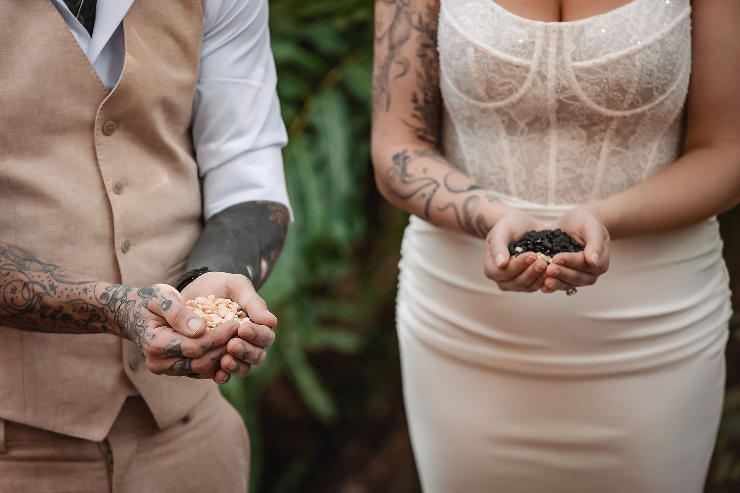 Tattooed couple holding handfuls of sand and dark stones during an outdoor wedding ceremony.