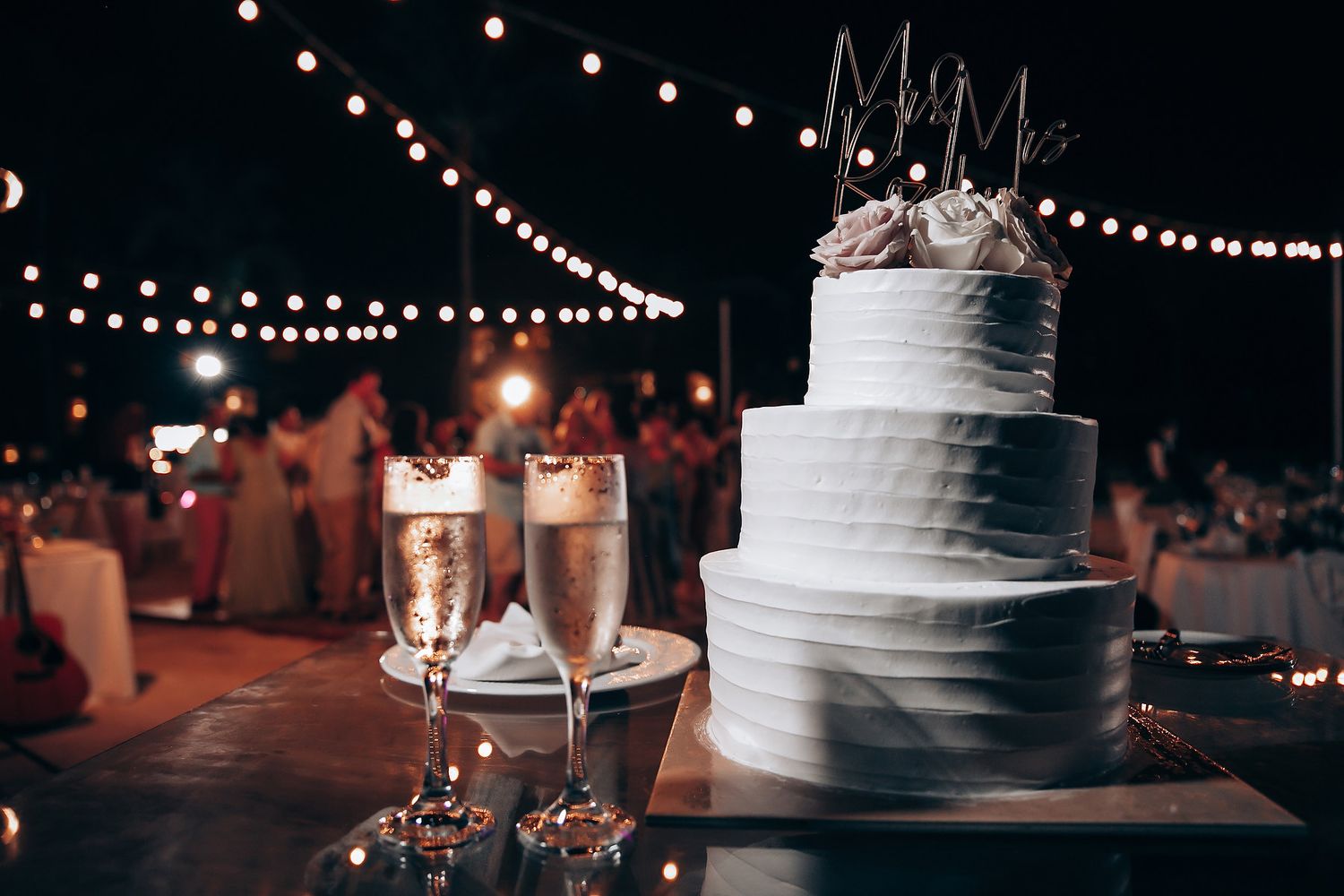 Wedding reception details with string lights and white tiered cake at night.