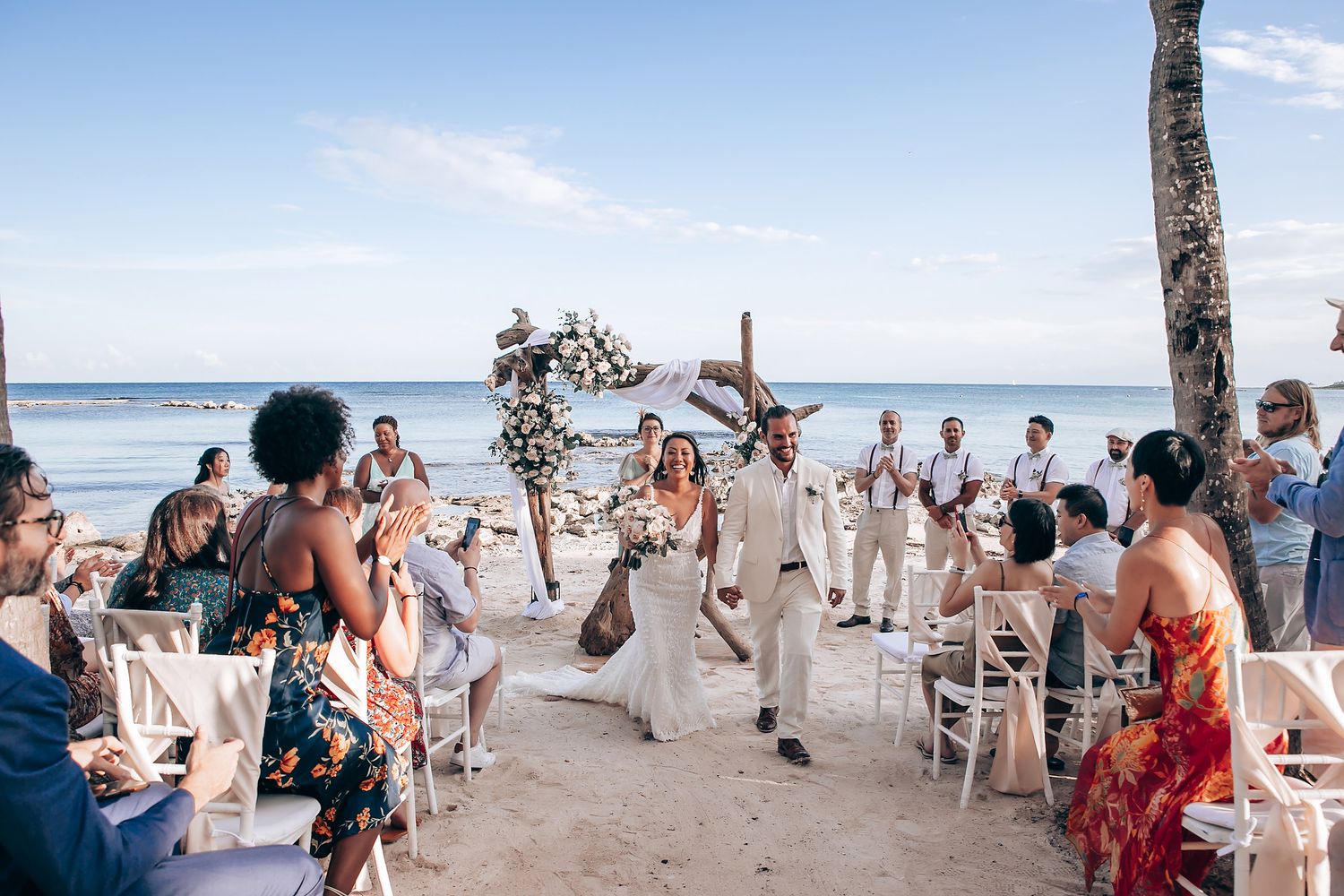 Beachfront wedding ceremony setup with white chairs and tropical palm trees.