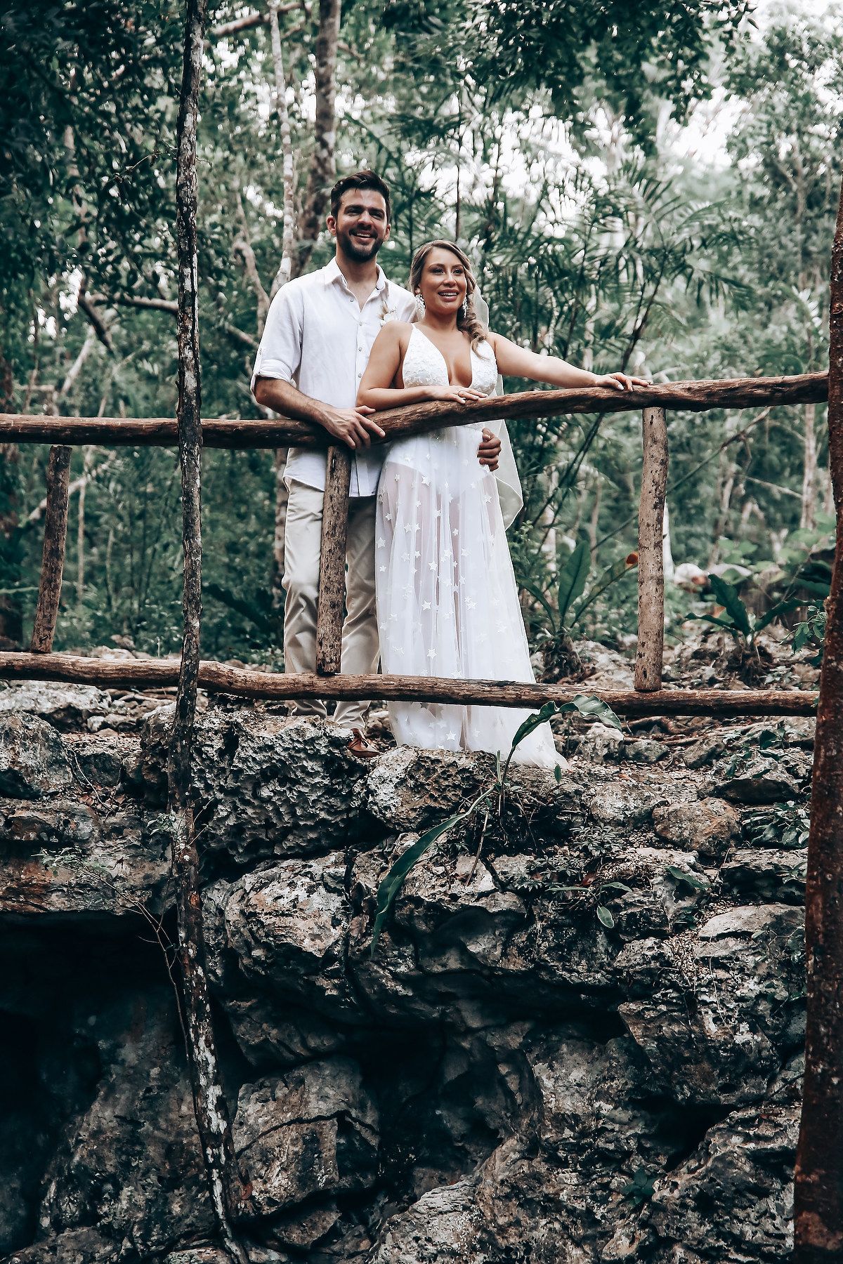 A couple stands on a rustic stone wall pathway surrounded by tropical forest vegetation.