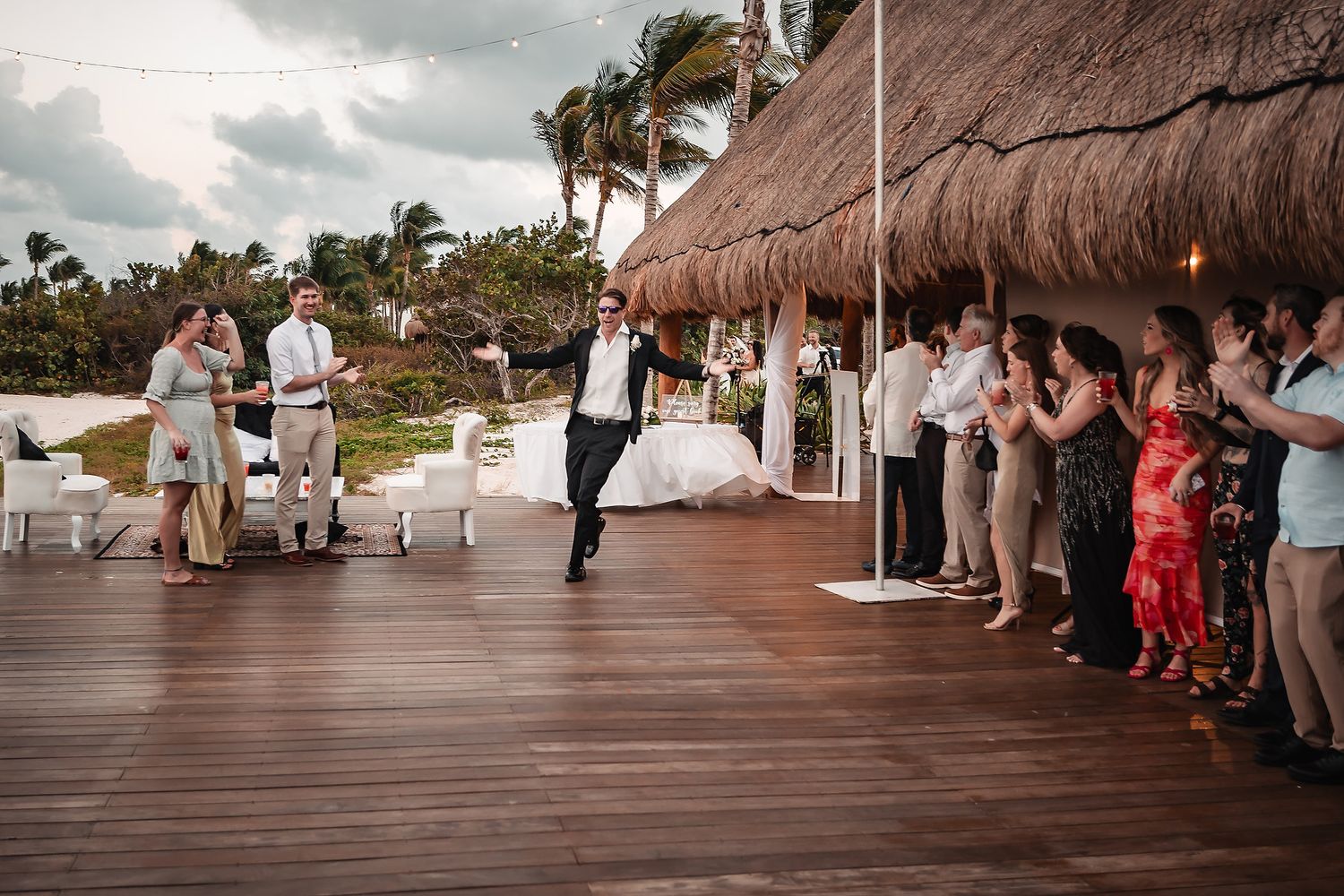 Groom dancing joyfully on wooden deck at tropical beach wedding with guests cheering under thatched roof.